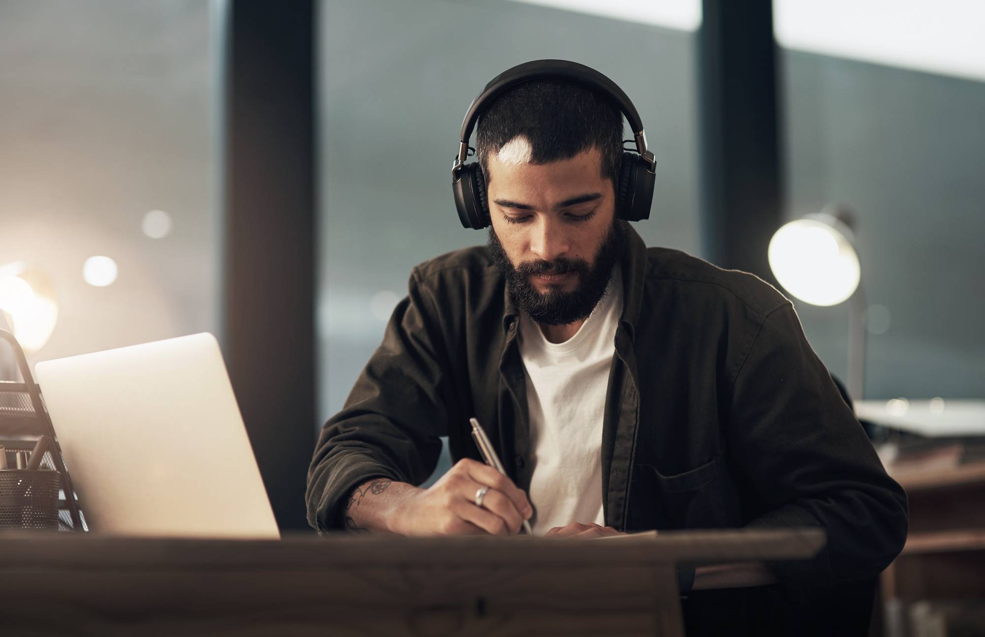 A man wearing headphones, writing in a notebook in front of his laptop