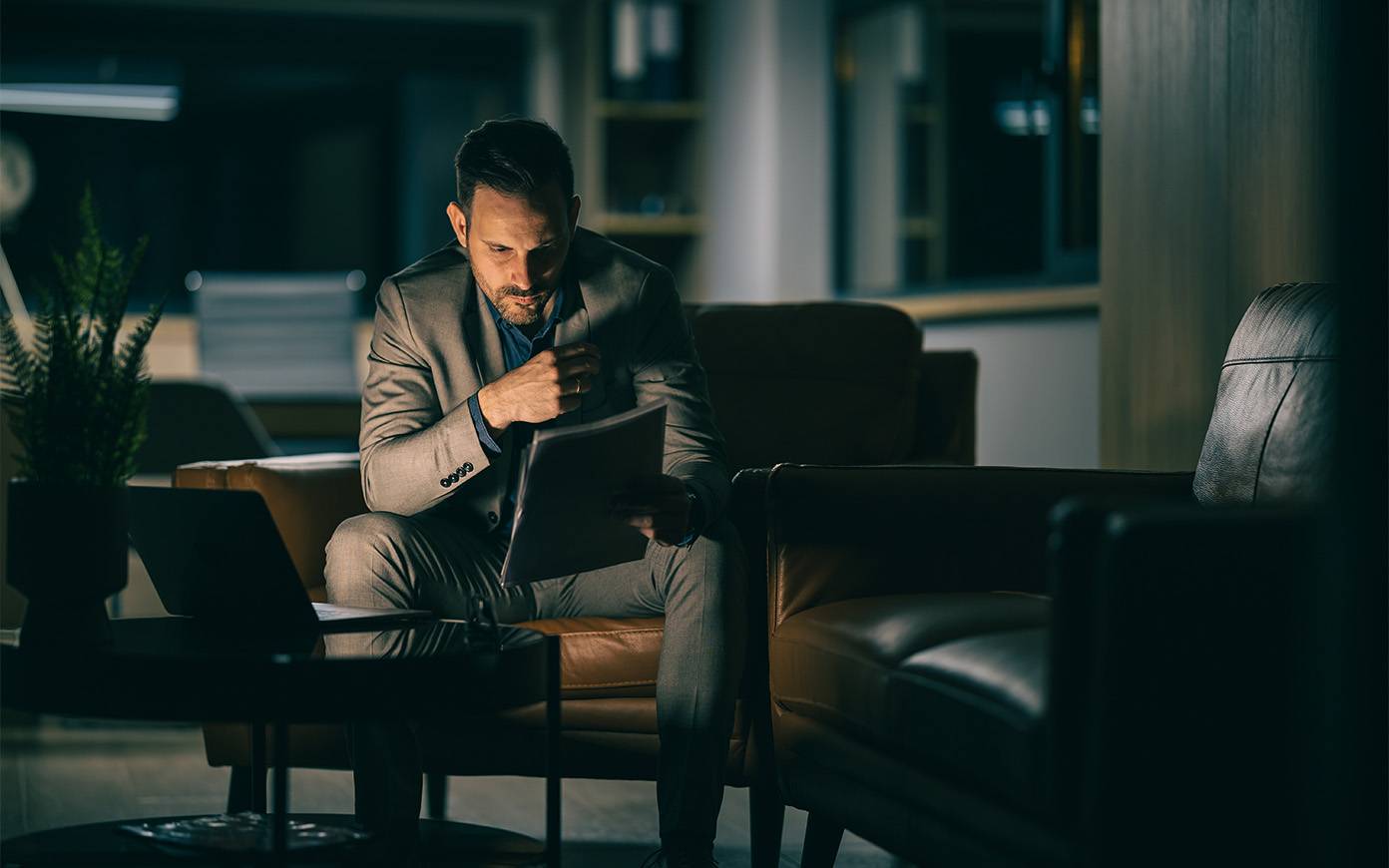 A man looking at a file in front of a laptop in a dark room